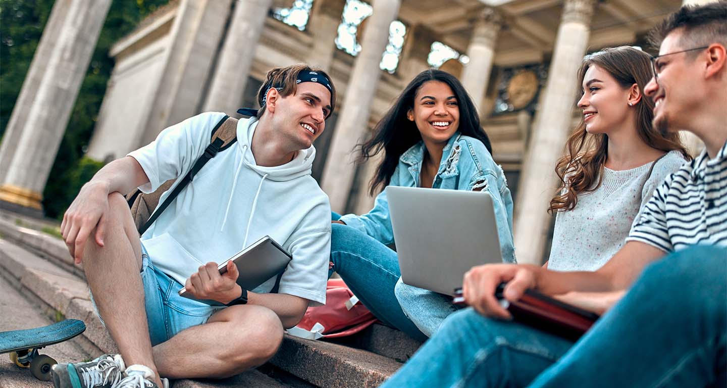 A group of students with laptops sit on the steps near the campus